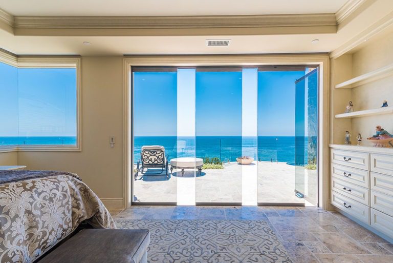 Looking at bedroom with stone flooring and shelving with staggered frameless glass doors with unobstructed view of the patio and ocean.