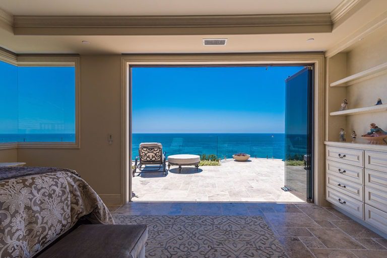 Looking at bedroom with stone flooring and shelving with stacked frameless glass doors stacked to the right of the door frame with view of the patio and ocean.