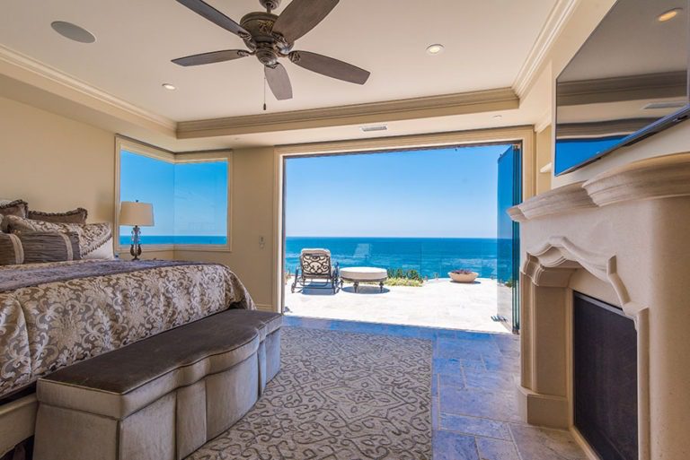 Looking at bedroom with stone flooring and stacked frameless glass doors stacked to the right of the door frame with view of the patio and ocean.