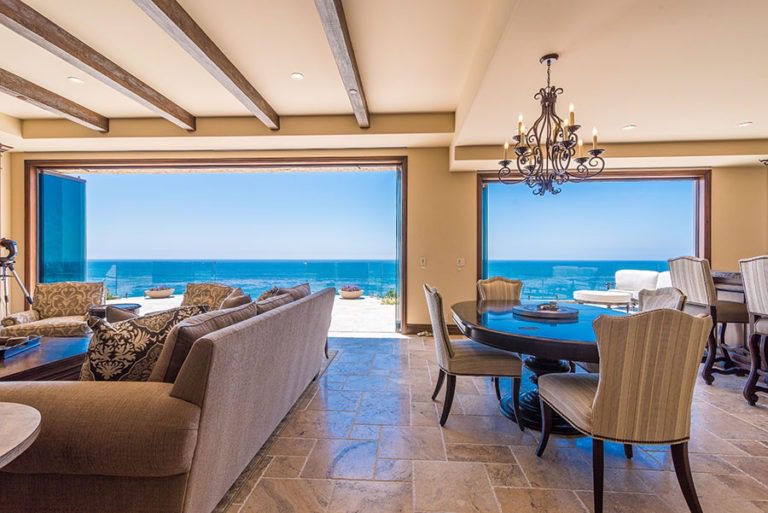 Open concept dining room and living room with stone floor and frameless glass doors stacked on sides of door frame with views of the ocean and patio.