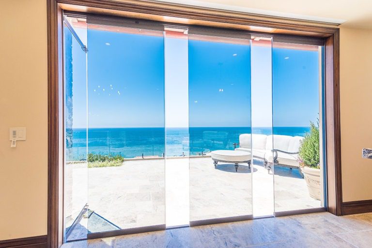 Looking at stone patio with patio furniture in corner with frameless sliding glass doors staggered in middle of door frame with one frame swung open allowing for unobstructed views of the ocean and patio.