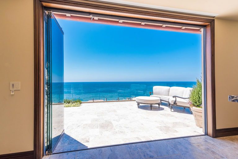 Looking at stone patio with patio furniture in corner with frameless sliding glass doors stacked on the right side of the door frame allowing for unobstructed views of the ocean and patio.