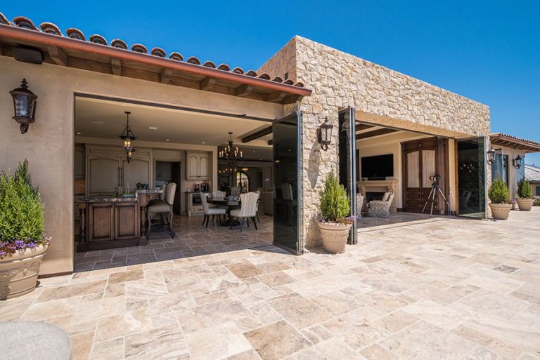 Looking from stone floor patio into open concept living, kitchen and dining room with frameless sliding glass doors stacked on the sides of the door frame.