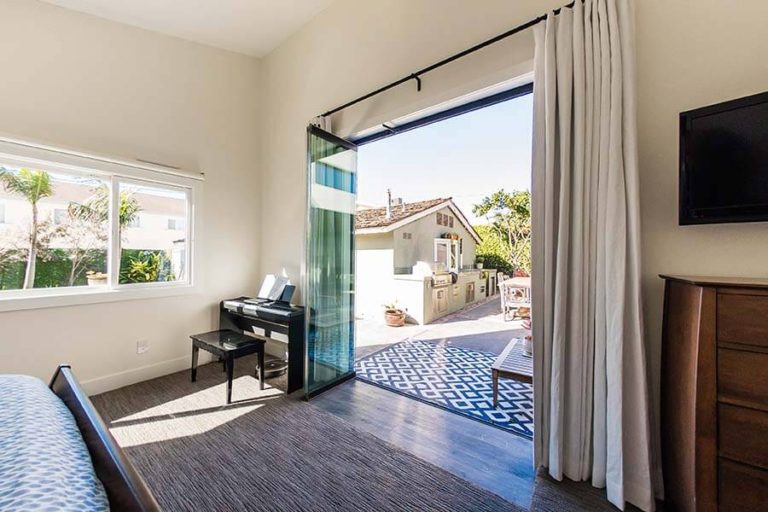 Bedroom with piano in the corner and stacked frameless sliding glass doors connecting to the outside area.