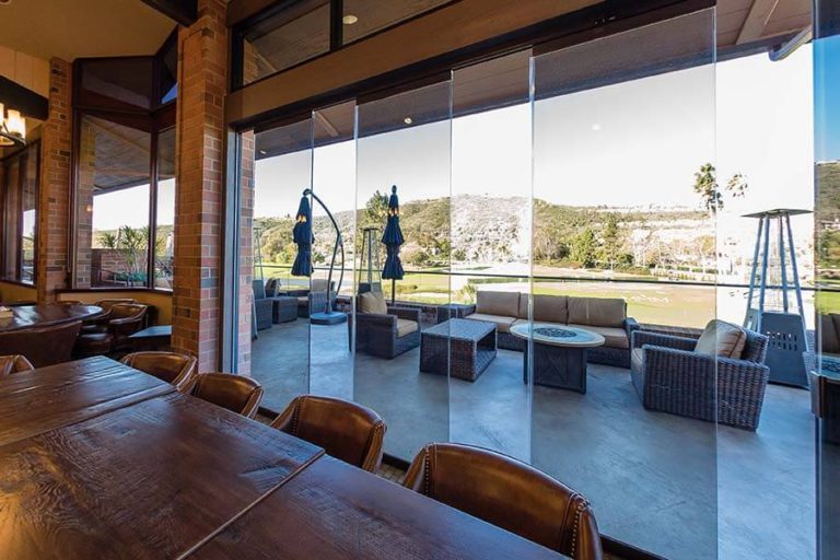 Country club with wood tables and leather chairs overlooking the yard through partially open frameless sliding glass doors.