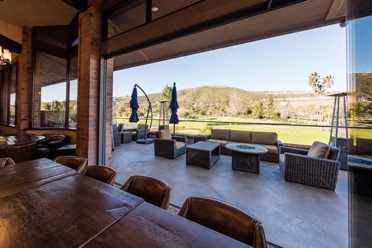 Country club with wood tables and leather chairs overlooking the yard through stacked frameless sliding glass doors.