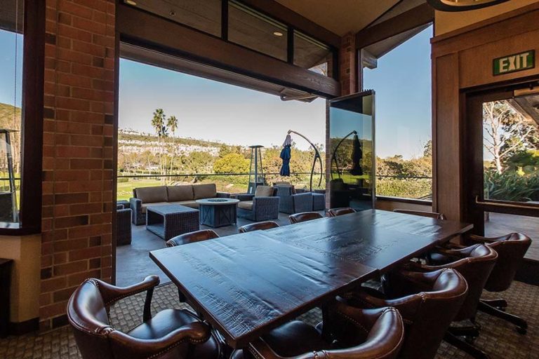 Country club with wood tables and leather chairs overlooking the patio and the yard through stacked frameless sliding glass doors.