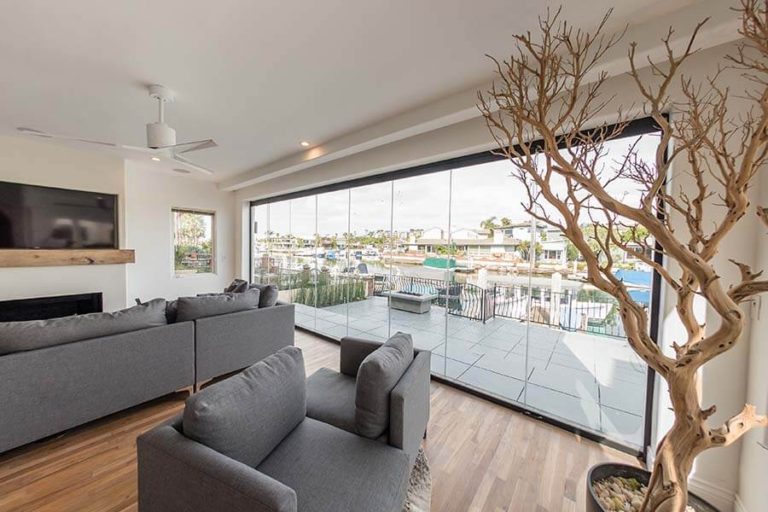 Living room with wood floor and grey couches overlooking outdoor area with closed frameless sliding glass doors.