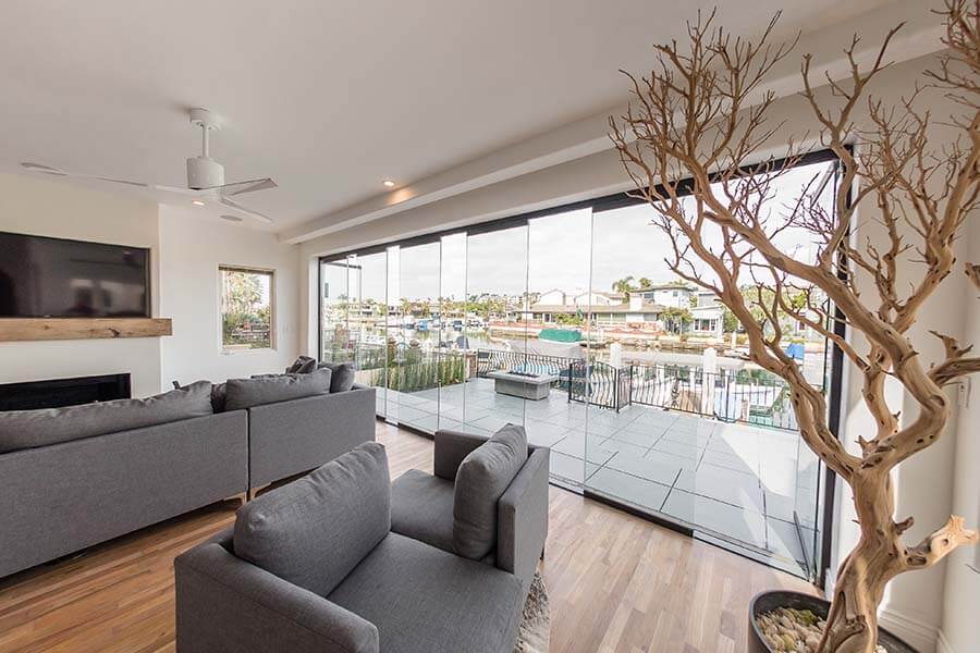 Living room with wood floor and grey couches overlooking outdoor area with partially open frameless sliding glass doors.