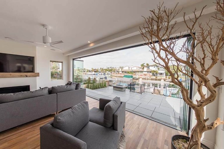 Living room with wood floor and grey couches overlooking outdoor area with stacked frameless sliding glass doors.