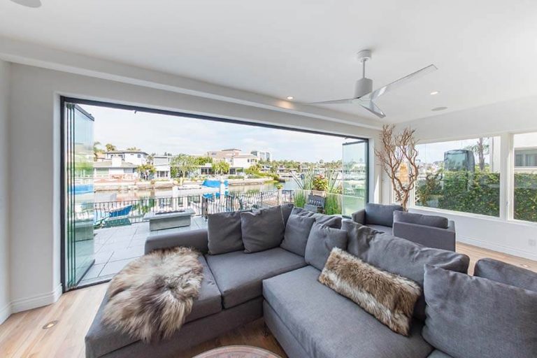 Living room with grey sectional sofa and wood floor overlooking outdoor patio through stacked frameless sliding glass doors.