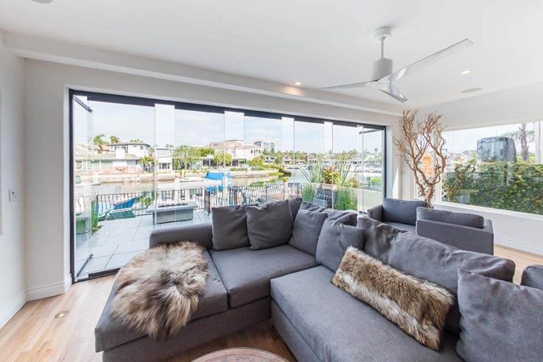 Living room with grey sectional sofa and wood floor overlooking outdoor patio through partially open frameless sliding glass doors.