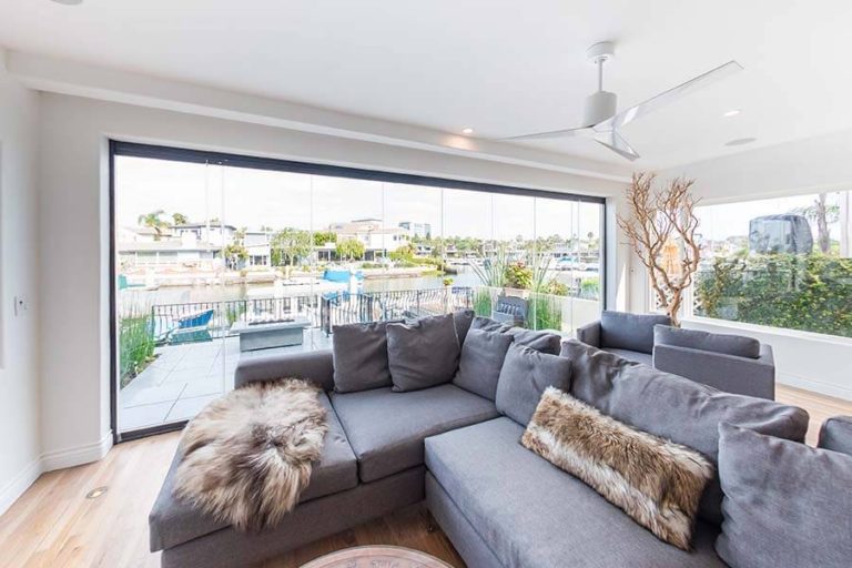 Living room with grey sectional sofa and wood floor overlooking outdoor patio through frameless sliding glass doors.