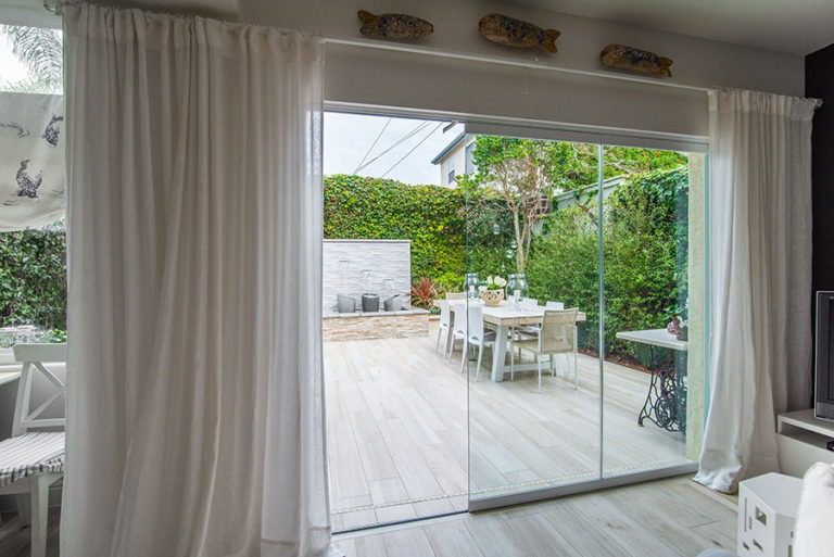 Enclosed frameless sliding glass doors with one frame swung open with an unobstructed view of backyard with fountain and table.