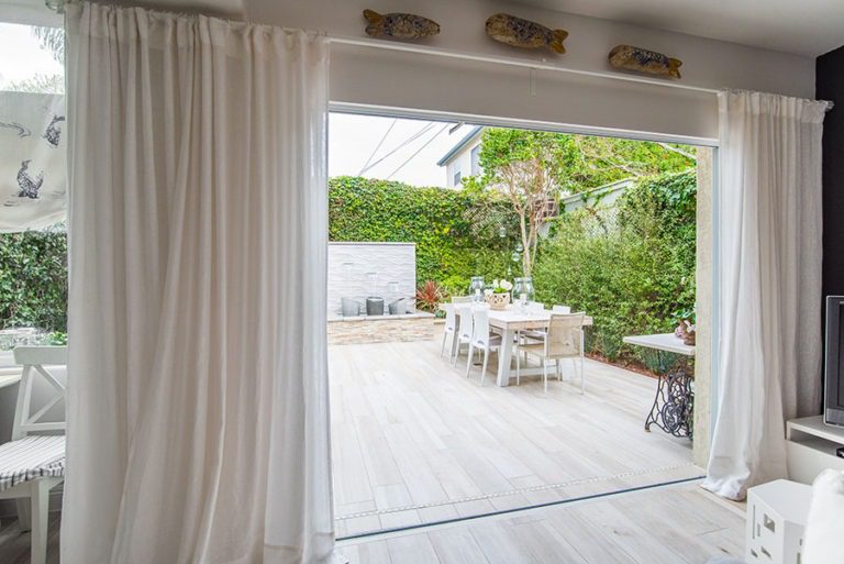 Stacked frameless sliding glass doors to the left of the doorframe with an unobstructed view of backyard with fountain and table.