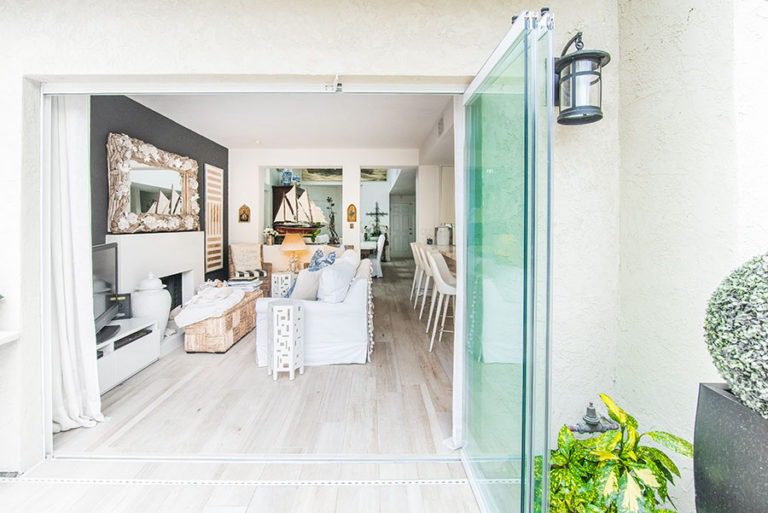 Looking into living room and kitchen with wood flooring from backyard with frameless sliding glass doors stacked to right side of door frame.