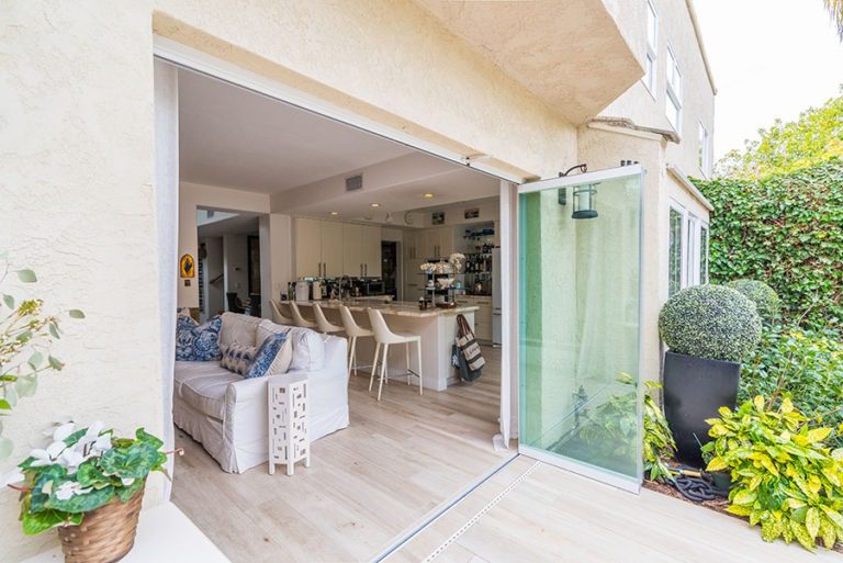 Looking into beautifully designed living room and kitchen with wood flooring from backyard with frameless sliding glass doors stacked to right side of door frame.