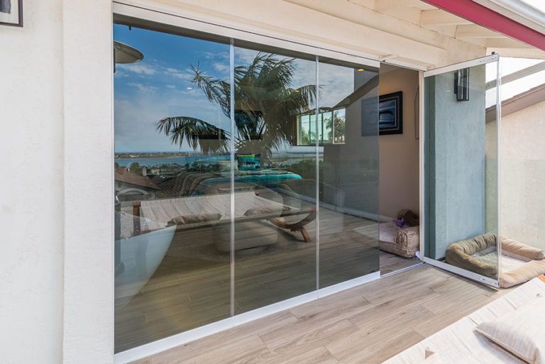 Looking from wooden floor patio at enclosed frameless sliding glass doors with one door frame swung open to the bedroom.