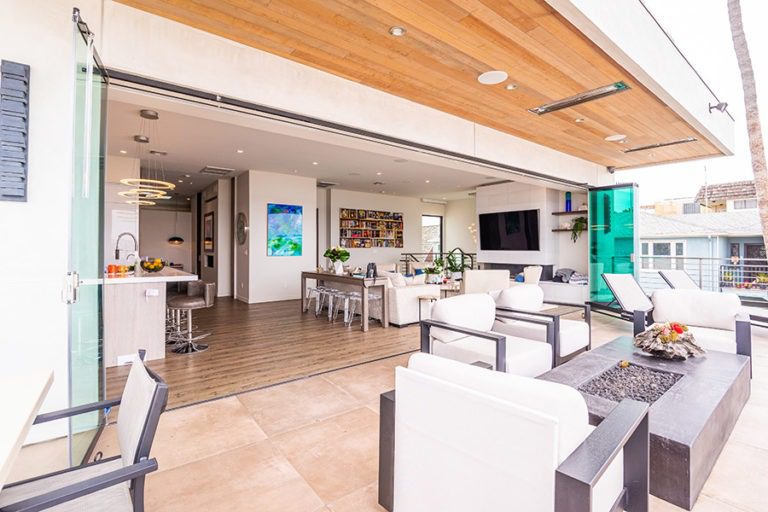 Marble floored patio with fire place looking into open concept of living room and kitchen with frameless glass doors stacked on each side of the door frame.