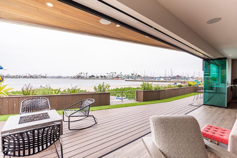 Frameless glass doors stacked to right side of room looking out to patio with wood floors and fire pit viewing the boat dock.