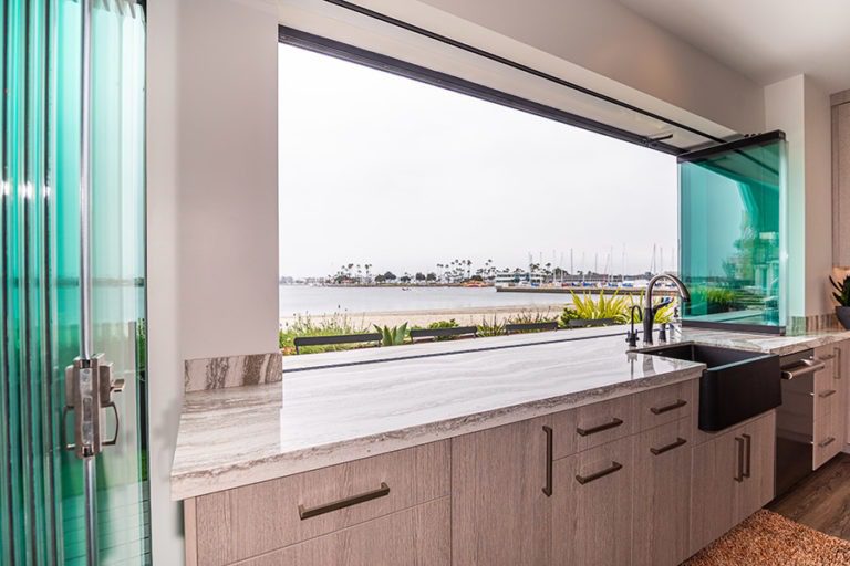 Marbled kitchen counter top with frameless glass doors and windows stacked to each side with unhindered views of the boat dock and the beach.