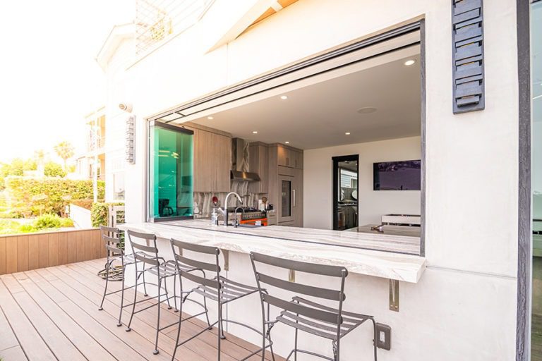 The sliding glass windows are stacked together on the left of the marble counter looking into the open concept kitchen from the wood floor patio.